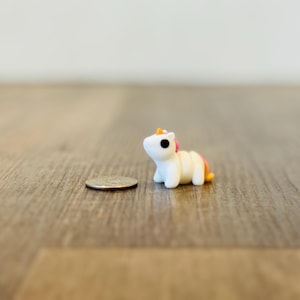 May include: A miniature white unicorn figurine with black eyes, a yellow horn, and pink and orange accents. The small, detailed sculpture is next to a quarter coin on a wooden surface.