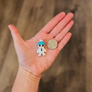 May include: A small, white snowman figurine with a blue hat and black button eyes and mouth. A coin is next to the snowman. The figurine is held in a person's hand.