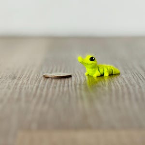 May include: A small, lime green alligator figurine with black eyes, lying on a wooden surface next to a quarter. The toy alligator is segmented and has a curved snout. The background is a blurred, light-colored wall.