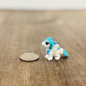 Może przedstawiać: Miniaturowa, biało-niebieska figurka smoka z monetą obok. Smok ma niebieskie detale na głowie, skrzydłach i ogonie. Moneta ma średnicę około 2,5 cm. Na drewnianej powierzchni.