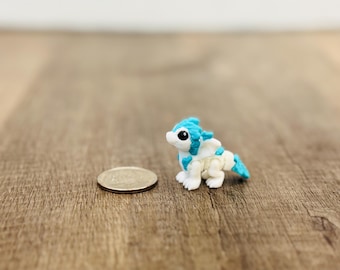 May include: A miniature, white and blue dragon figurine with a coin next to it. The dragon has a detailed design with blue accents on its head, wings, and tail. The coin is a US quarter, approximately 1 inch in diameter.