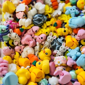 May include: A vibrant assortment of colorful polymer clay charms. The collection includes miniature ducks, bunnies, carrots, bees, and other whimsical shapes in shades of yellow, pink, orange, blue, and white. These charms are ideal for crafting and jewelry making.