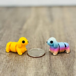May include: Two miniature dachshund dog figurines. One is yellow, the other is blue, pink, and purple. A quarter is placed between them for size comparison. The dogs are segmented, with each section of the body able to move.