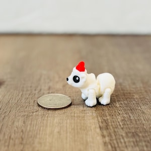 May include: A small, white polar bear figurine with a red Santa hat, black eyes, and a quarter coin next to it. The figurine is on a wooden surface.