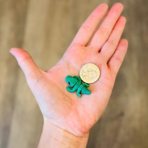 May include: A small, teal-colored alligator figurine with detailed texture, next to a quarter coin. The alligator is lying on its belly with its legs outstretched. The coin is approximately 1 inch in diameter. The figurine is likely a toy or decorative item.