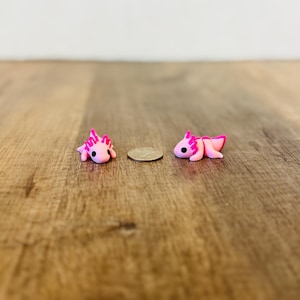 May include: Two small, pink axolotl figurines with bright pink gills and accents are displayed on a wooden surface. A coin is placed between the figurines for size comparison. The axolotls are lying down, with their legs and tails curled.