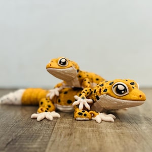 May include: Two detailed gecko figurines. The geckos are yellow with black spots and white feet. One gecko is on top of the other. The geckos have large, expressive eyes and are posed on a wooden surface.