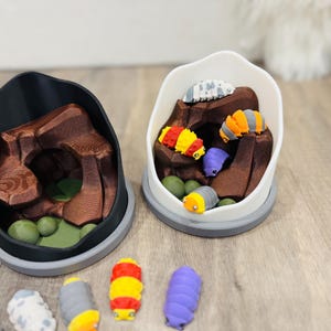 May include: Two 3D-printed terrariums, one black and one white, each containing a brown rock formation and colorful, segmented caterpillar figurines. The caterpillars are in various colors, including red, yellow, orange, and purple.