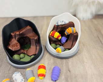 May include: Two 3D-printed terrariums, one black and one white, each containing a brown rock formation and colourful, segmented caterpillar figurines. The caterpillars are in various colours, including red, yellow, orange, and purple.