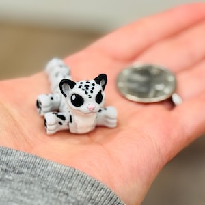 May include: A miniature white and black spotted leopard figurine with large black eyes and a pink nose. The figurine is lying down on a hand next to a coin. The leopard is a cute, small collectible.