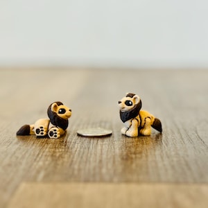 May include: Two miniature lion figurines, each with a black mane and tail, tan bodies, and black paws. They are positioned on a wooden surface, with a coin between them. The figurines are detailed and appear handcrafted.