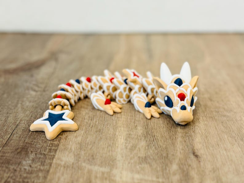 Pu&ograve; includere: Statuetta di drago articolata beige con dettagli bianchi, rossi e blu. Il drago ha una coda a forma di stella. La statuetta &egrave; realizzata in un materiale di colore chiaro e poggia su una superficie di legno.