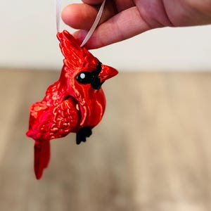 May include: A vibrant red cardinal ornament with black accents and a white ribbon for hanging. The bird has detailed feather textures and a glossy finish, perfect for holiday or nature-themed decor.
