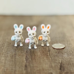 Pode incluir: Três pequenas figuras de esqueleto branco com orelhas de coelho e segurando ovos coloridos. As orelhas de coelho são azuis, rosa e laranja. Uma moeda está ao lado das figuras em uma superfície de madeira.