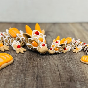 May include: A pair of articulated dragon sculptures. The dragons are tan with white, orange, and red accents. The larger dragon has a detailed head and body, while the smaller dragon has a similar design. The dragons are on a wooden surface.