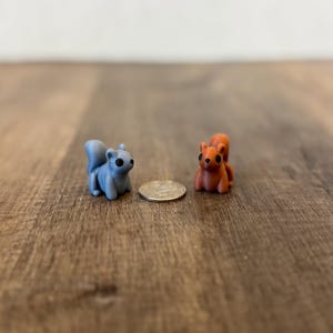May include: Two miniature squirrel figurines, one blue and one orange, sit on a wooden surface. The squirrels are next to a coin, providing a size reference. The figurines have detailed features and dark eyes.