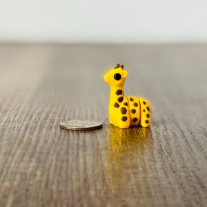 May include: A small, yellow giraffe figurine with black spots and a brown mane. The miniature animal is next to a quarter coin, suggesting its small size. The giraffe is on a wooden surface.