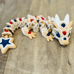 Pu&ograve; includere: Una figura di drago articolata in beige, bianco, rosso e blu. Il drago ha una coda a forma di stella ed &egrave; posizionato su una superficie di legno. Il design incorpora colori patriottici e un'estetica giocosa.