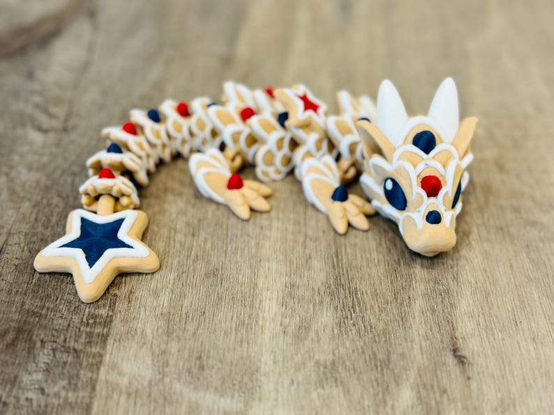 Pu&ograve; includere: Una figura di drago articolata in beige, bianco, rosso e blu. Il drago ha una coda a forma di stella ed &egrave; posizionato su una superficie di legno. Il design incorpora colori patriottici e un'estetica giocosa.