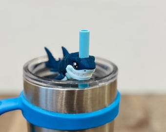 May include: Stainless steel tumbler with a blue handle and a blue shark-shaped straw topper. The tumbler has a clear lid and a blue silicone band around the middle. The shark has white teeth and eyes.