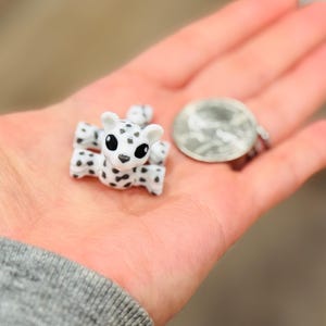 May include: A small, white and black spotted leopard figurine with large black eyes. The figurine is held in an open hand next to a quarter coin. The leopard has four legs and two small ears. The background is blurred.