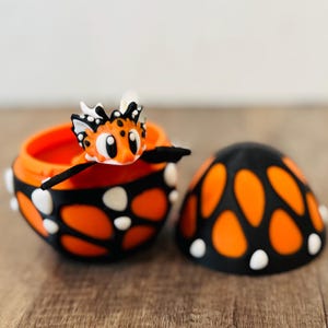 Op de afbeelding: Een oranje en zwarte decoratieve sieradendoos met een bijpassende deksel. De doos heeft een vlinderontwerp met witte accenten. Een kleine, cartoonachtige vlinderfiguur zit op de rand van de doos.