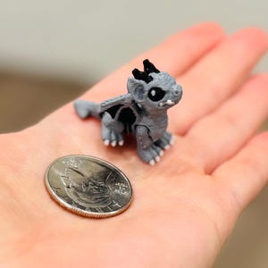 May include: A miniature gray dragon figurine with black accents on its head and wings, featuring white claws and large dark eyes. The small creature is positioned next to a silver United States quarter coin for scale, highlighting its tiny size.