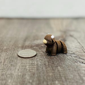 May include: A small, brown figurine of a Highland cow with white horns and a tan snout. The cow is segmented, and a quarter coin is next to it for scale. The figurine is on a wooden surface.