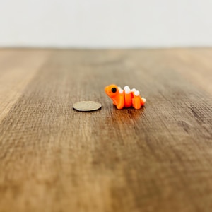 May include: A miniature orange and white polymer clay salamander figurine with black eyes. The figurine is next to a quarter coin on a wooden surface. The salamander is a handcrafted item.