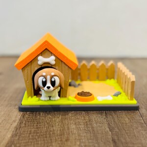 Puede incluir: Una caprichosa figura de caseta para perros con un beagle de dibujos animados asomándose. El techo naranja tiene un adorno de hueso. Una pequeña valla y un cuenco de comida completan la escena, todo sobre una base verde.