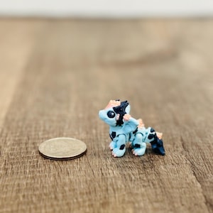 May include: A small, handcrafted dragon figurine in shades of light blue, black, and peach. The miniature sculpture is next to a quarter coin for size comparison. The dragon has detailed features and is posed on a wooden surface.
