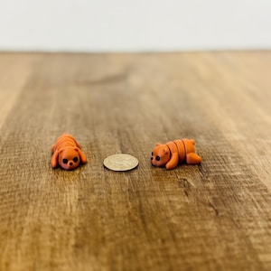 May include: Two small orange animal figurines, one resembling a dog and the other a bear, lie on a wooden surface. A coin is placed between them. The dog figurine is lying down, while the bear is in a resting position.