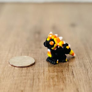 May include: A small black dragon figurine with orange, yellow, and white candy corn-shaped spikes along its back and tail. The dragon has large orange eyes and is positioned next to a silver United States quarter coin for scale reference.