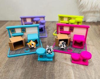 May include: Colorful cat-themed playsets with miniature cat figurines. Each set features a house, a box, a food bowl, and a cat. The sets are in various colors, including blue, purple, pink, and green. The cats have different fur patterns.