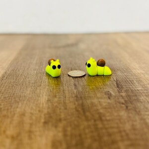May include: Two small, handmade polymer clay snail figurines. The snails are bright yellow with brown shell details and black eyes. A coin is placed between the snails. The figurines are on a wooden surface.