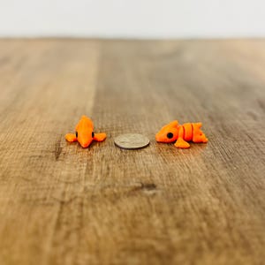 May include: Two small, orange, fish-shaped figurines with black eyes are displayed on a wooden surface. A U.S. quarter is placed between the figurines. The figurines are approximately 1 inch in length. The background is a light brown wooden surface.
