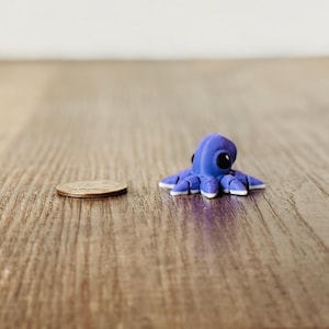 May include: A small, blue octopus figurine with white-tipped tentacles and large black eyes. The figurine is next to a U.S. quarter coin on a wooden surface. The octopus is approximately 1 inch in diameter.