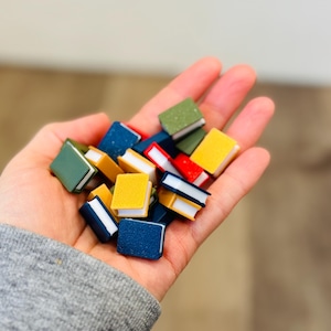 May include: A hand holding a collection of miniature books. The books have colorful covers in shades of green, yellow, blue, and red. The pages are white, and the books are small, likely for a dollhouse or craft project.