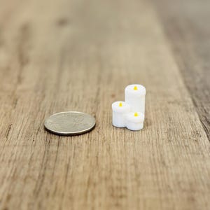May include: Miniature white candles with yellow flames are grouped together on a wooden surface. A coin is next to the candles for size comparison. The candles are of varying heights and shapes, creating a cozy, decorative display.