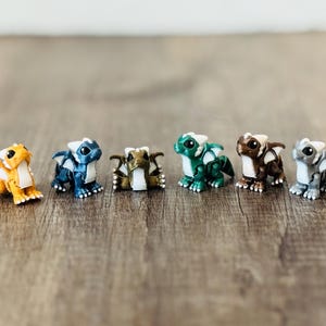 May include: A set of six small dragon figurines arranged in a row. The dragons are colored orange, blue, gold, green, brown, and gray. Each figurine features white accents on the wings, horns, and chest, with large black eyes and a seated posture.