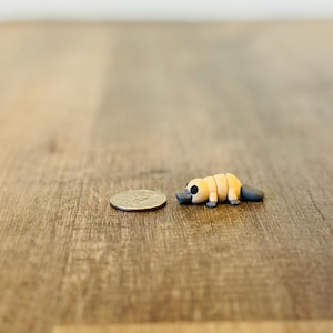 Può includere: Una statuetta di ornitorinco in miniatura fatta a mano, con un corpo beige, un design segmentato e un becco e una coda grigio scuro. La statuetta è accanto a una moneta, che mostra le sue piccole dimensioni. Lo sfondo è una superficie di legno.