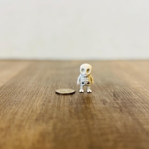 Può includere: Una piccola statuina di scheletro bianco è accanto a una moneta su una superficie di legno. La statuina ha una gabbia toracica dettagliata e orbite oculari vuote. La moneta è di colore argento.