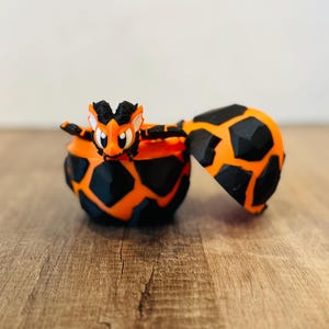 May include: A 3D-printed orange and black dragon figurine emerges from a matching egg-shaped container. The dragon has a detailed design with outstretched arms. The container is open, revealing the interior.