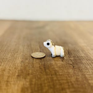May include: A small, white and tan toy horse figurine with black eyes and a gray mane. The miniature horse is positioned next to a quarter coin on a wooden surface. The figurine is a collectible item.