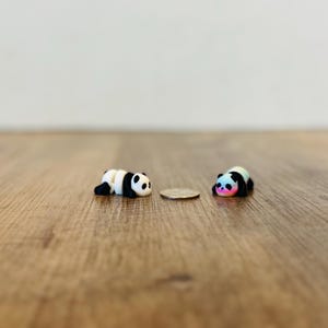 May include: Two miniature panda figurines, one with classic black and white markings, the other with a colourful pastel gradient. They are lying down on a wooden surface, next to a coin. The pandas are small, likely under 2.5 cm in length.
