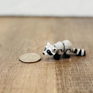 May include: A small, detailed figurine of a raccoon with a black and white striped tail, black paws, and a coin next to it. The raccoon is gray and white, with a black mask around its eyes. The figurine is on a wooden surface.