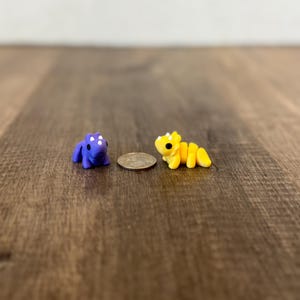 May include: A miniature purple dinosaur figurine with white spots and a yellow segmented dragon figurine are displayed next to a coin. The figurines are small, detailed, and likely handmade.