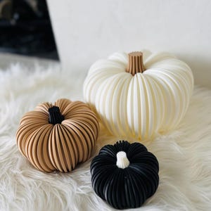 May include: Three decorative pumpkins in varying sizes and colors. The largest is white with a brown stem, the medium-sized pumpkin is tan with a black stem, and the smallest is black with a white stem. All have a ribbed design.
