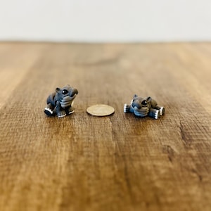 May include: Two small, gray figurines of hippos with white accents on their feet and a coin are displayed on a wooden surface. One hippo is upright, the other is lying down. The background is a plain white wall.