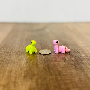 May include: Two small, colorful clay dinosaur figurines. One is lime green, the other is pink. They are positioned on a wooden surface with a coin between them. The dinosaurs have black eyes and are in a playful pose.